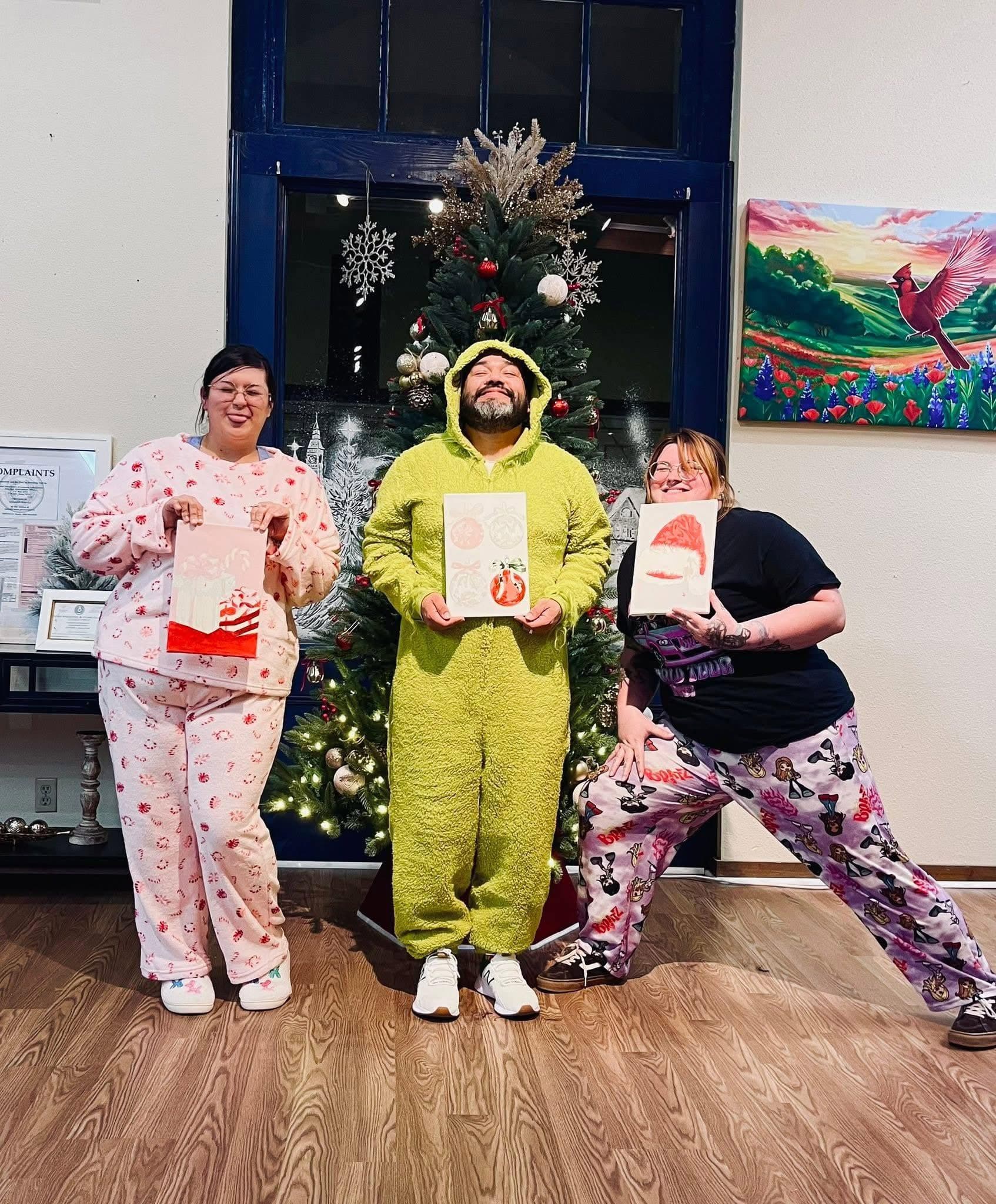 Three people in holiday pajamas holding small paintings in front of a Christmas tree at the studio.