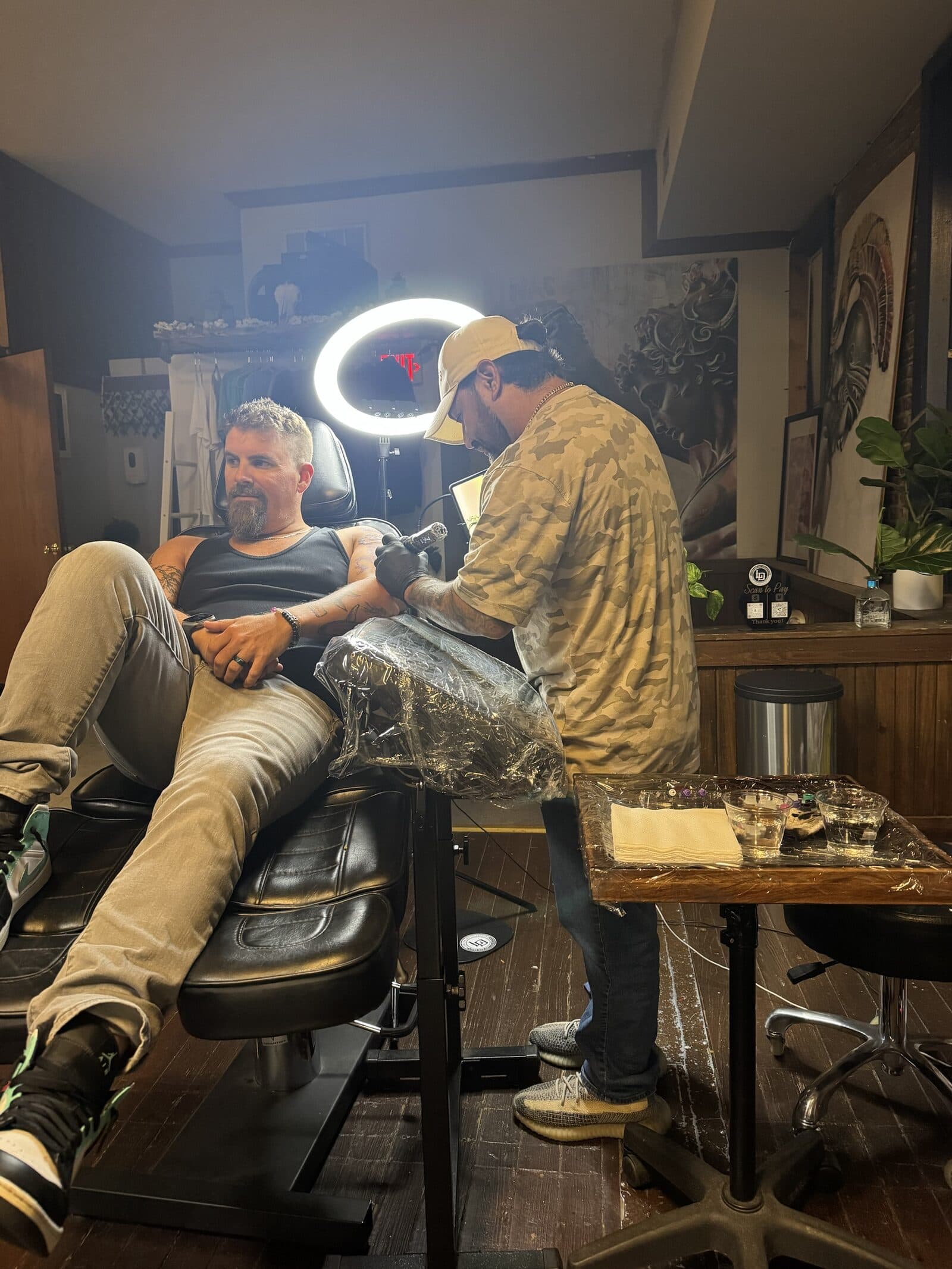 Jeremiah tattooing a client’s arm under a ring light, with his large paintings and a fiddle-leaf fig behind them.