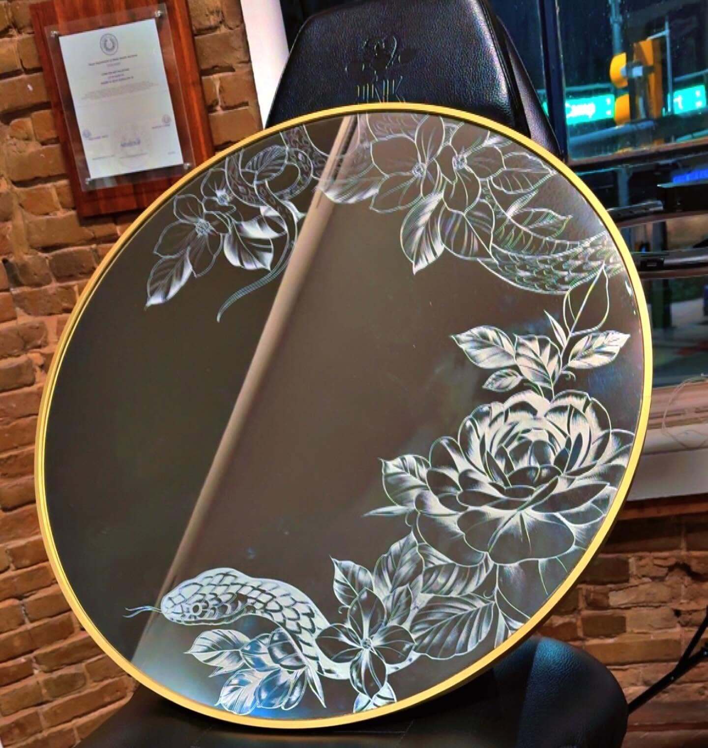A round gold-framed mirror etched with roses and leaves, resting on a tattoo chair.