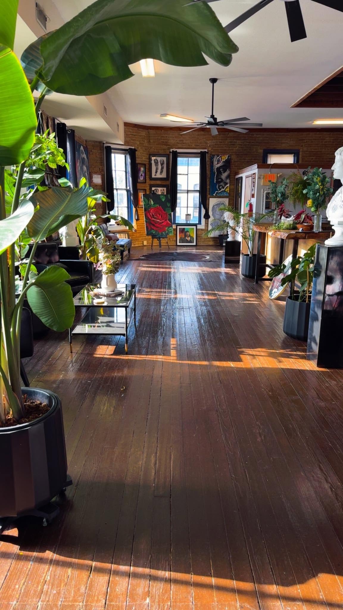 Inside Lions Den: afternoon sun across wood floors, tall plants, brick walls hung with paintings.